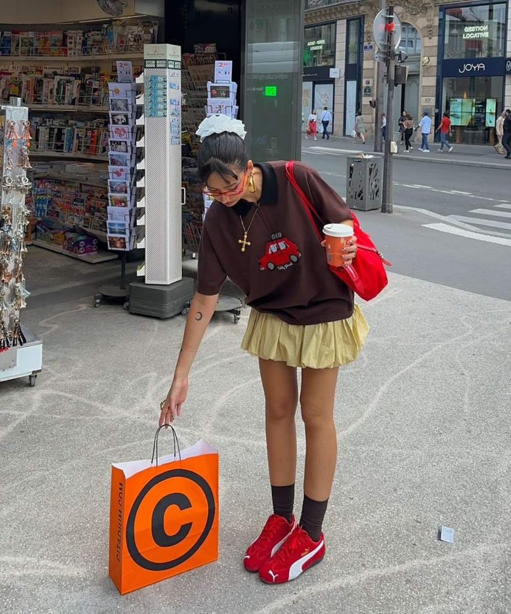 Jovem vestindo camiseta oversized marron com estampa, saia curta plissada em tom pastel, tênis vermelhos e meias pretas. Acessórios incluem scrunchie branco no cabelo, bolsa de ombro vermelha e óculos escuros. Estilo urbano descontraído e moderno.