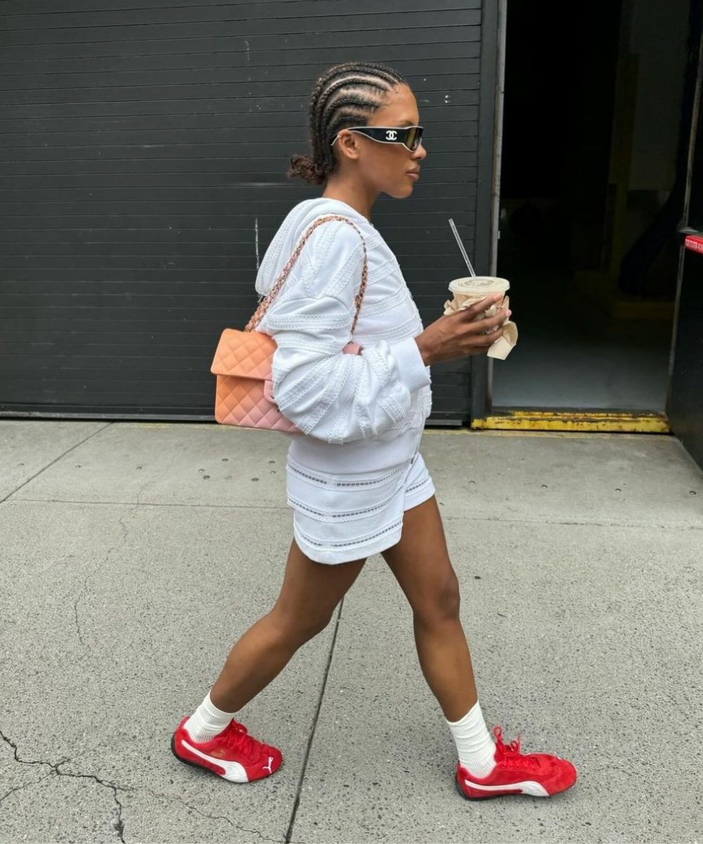 Moda enquanto caminha: mulher com tranças, conjunto branco de tecido texturizado, tênis vermelhos, meias brancas e bolsa acolchoada bege com corrente. Óculos de sol de marca fazem parte do estilo urbano e despojado.