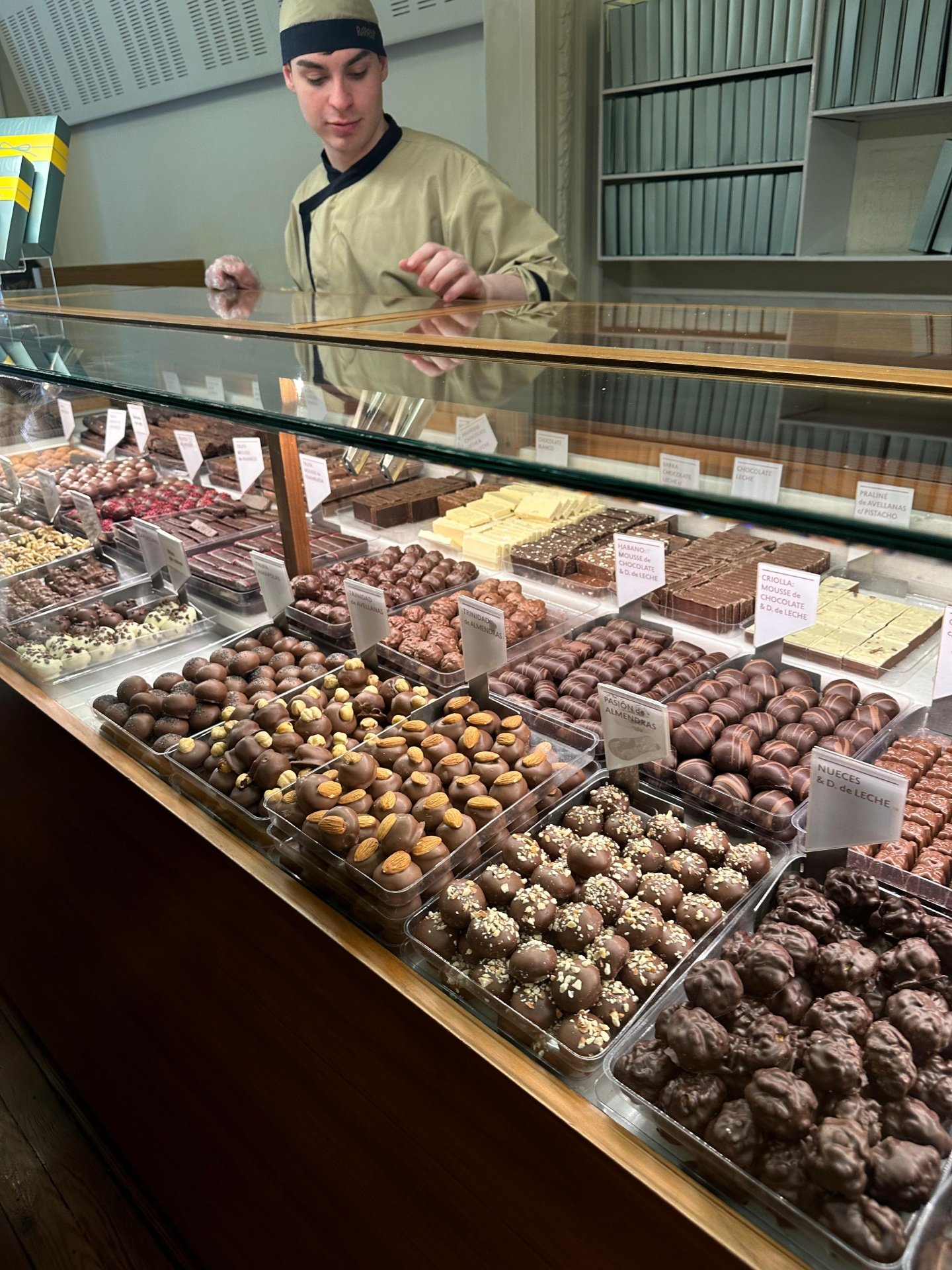 Vendedor de chocolates usando uniforme bege com detalhes pretos, composto por jaqueta e touca de chef. Ambiente com prateleiras cheias de chocolates arrumados em bandejas. Restaurantes em Buenos Aires