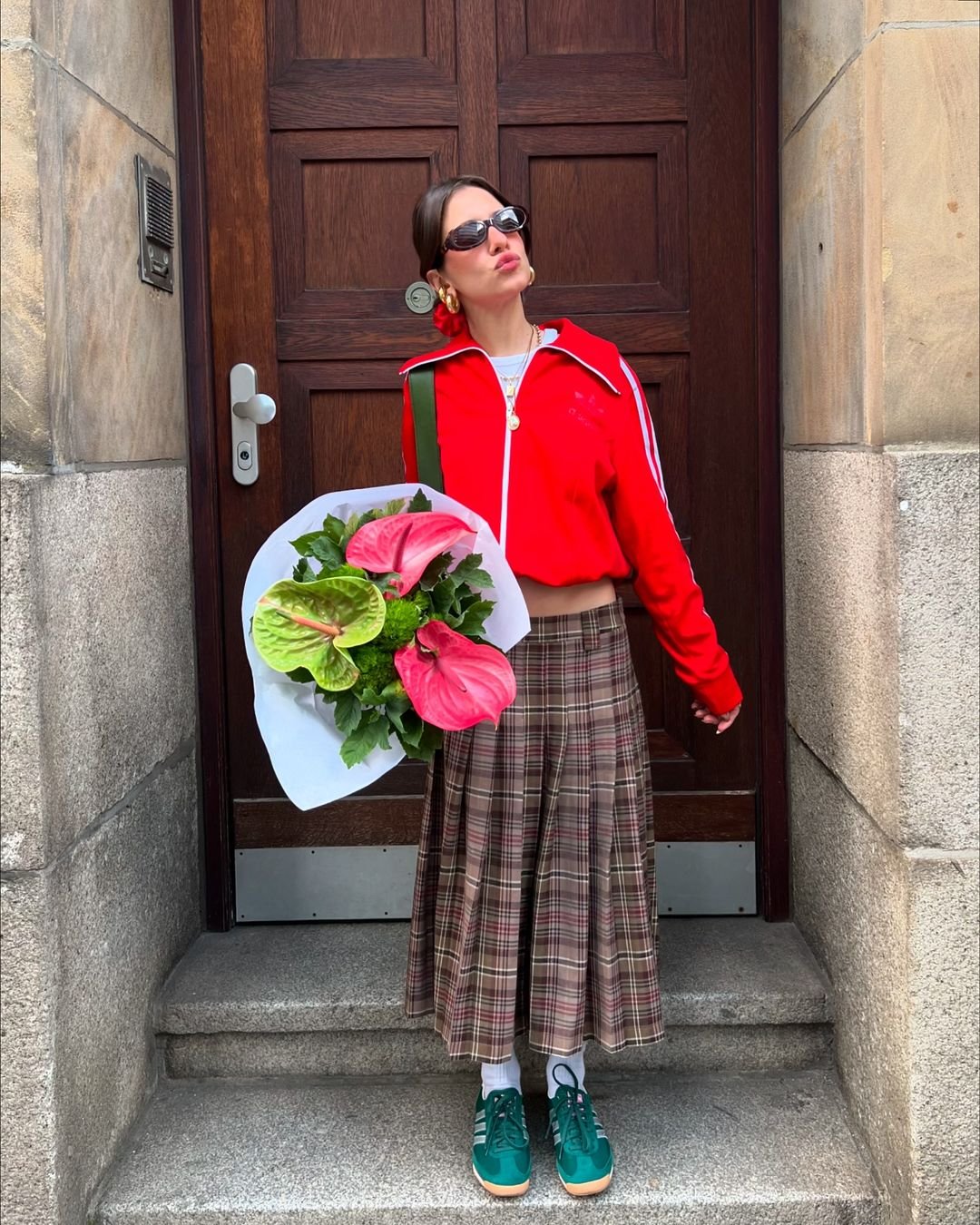 Mulher com look urbano e casual, usando jaqueta vermelha com zíper, óculos de sol preto, saia xadrez vintage, tênis verde com meias brancas e brincos dourados grandes. Ela carrega um buquê de flores grandes coloridas.