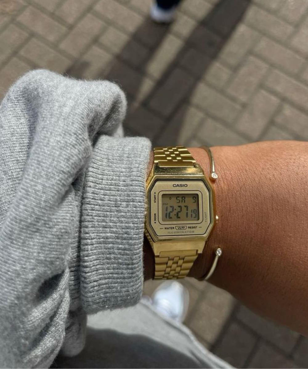 Imagem mostra um pulso com relógio digital Casio dourado e uma pulseira simples. Suéter cinza e calça combinando estão visíveis, enfatizando um estilo casual e retrô. A tendência de moda mistura conforto com acessórios vintage.