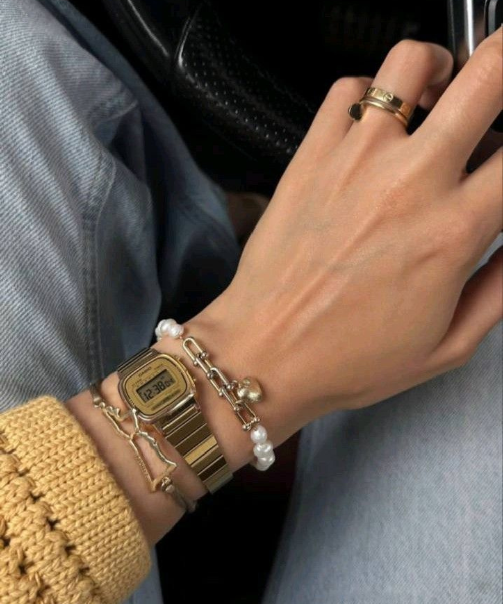 Imagem de um pulso usando relógio digital dourado estilo retrô, braceletes delicados com pérolas e anéis dourados minimalistas. Moda casual com suéter tricotado bege e calça jeans. Tendências de acessórios elegantes e retrô integradas ao visual do dia a dia.