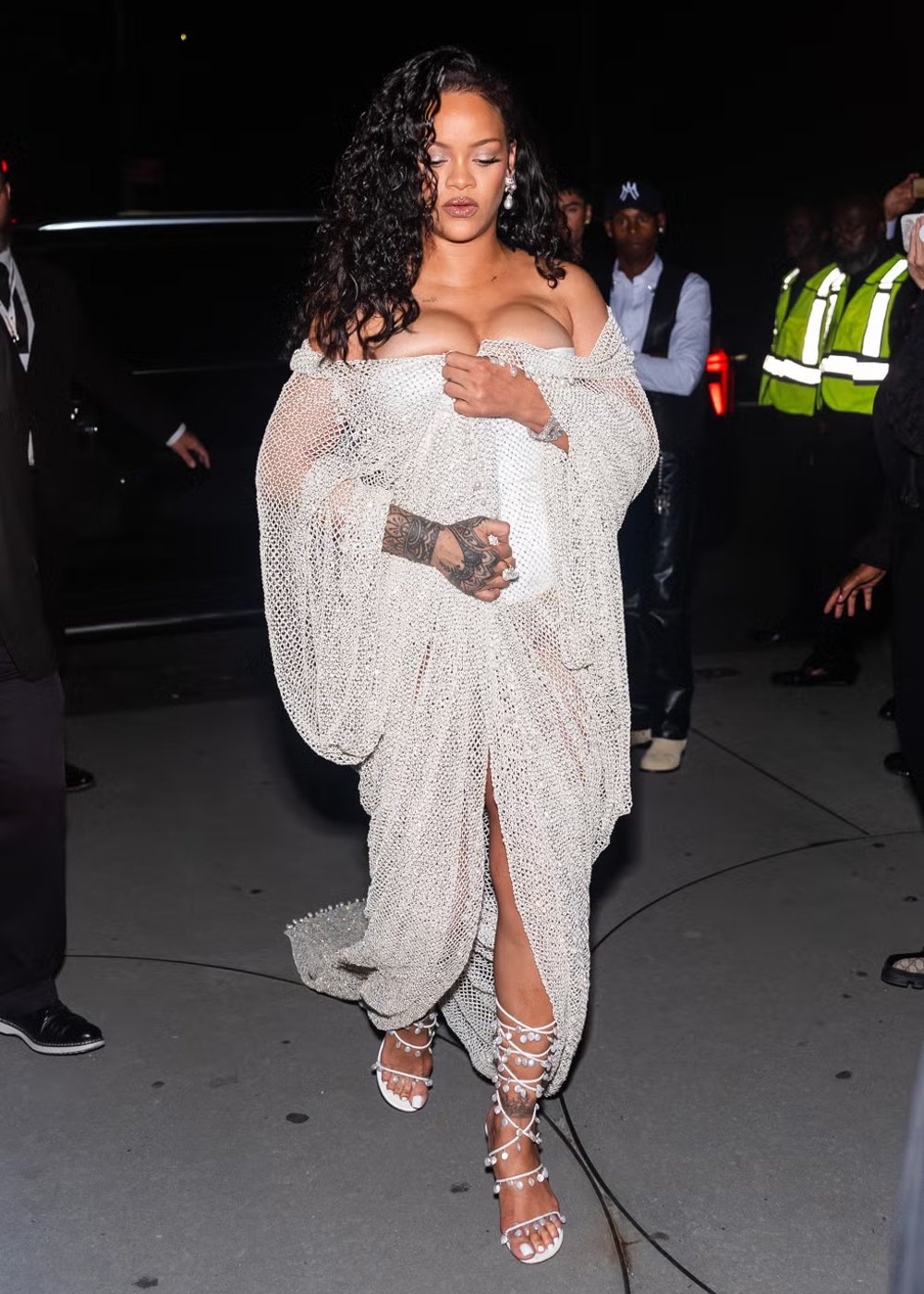 Rihanna usando vestido branco texturizado, ombros à mostra com decote ousado. Acessórios incluem sandálias brancas de tiras entrelaçadas. Look ousado e elegante. NYFW SS25