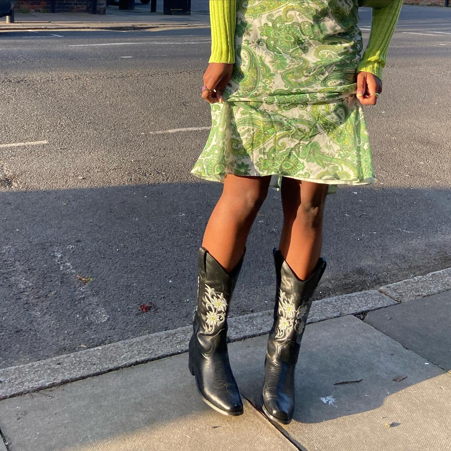 Pessoa vestindo uma saia verde estampada com arabescos, combinada com uma blusa de manga longa em tom neon. Completa o look com botas cowboy pretas, criando um visual moderno com influência boho-chic e western, destacando-se pelas cores vibrantes e padrão ousado.