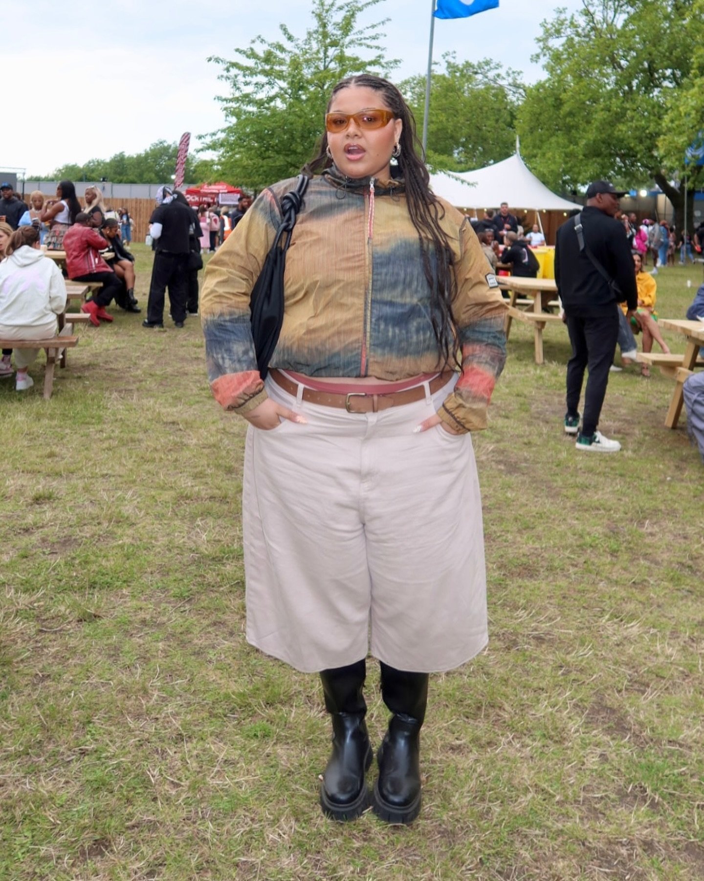 Pessoa em um evento ao ar livre, vestindo jaqueta tie-dye nos tons de marrom e azul, bermuda beige e botas pretas robustas. Completa o look com óculos de sol laranja e brincos grandes, refletindo uma mistura de tendências casuais e alternativas.