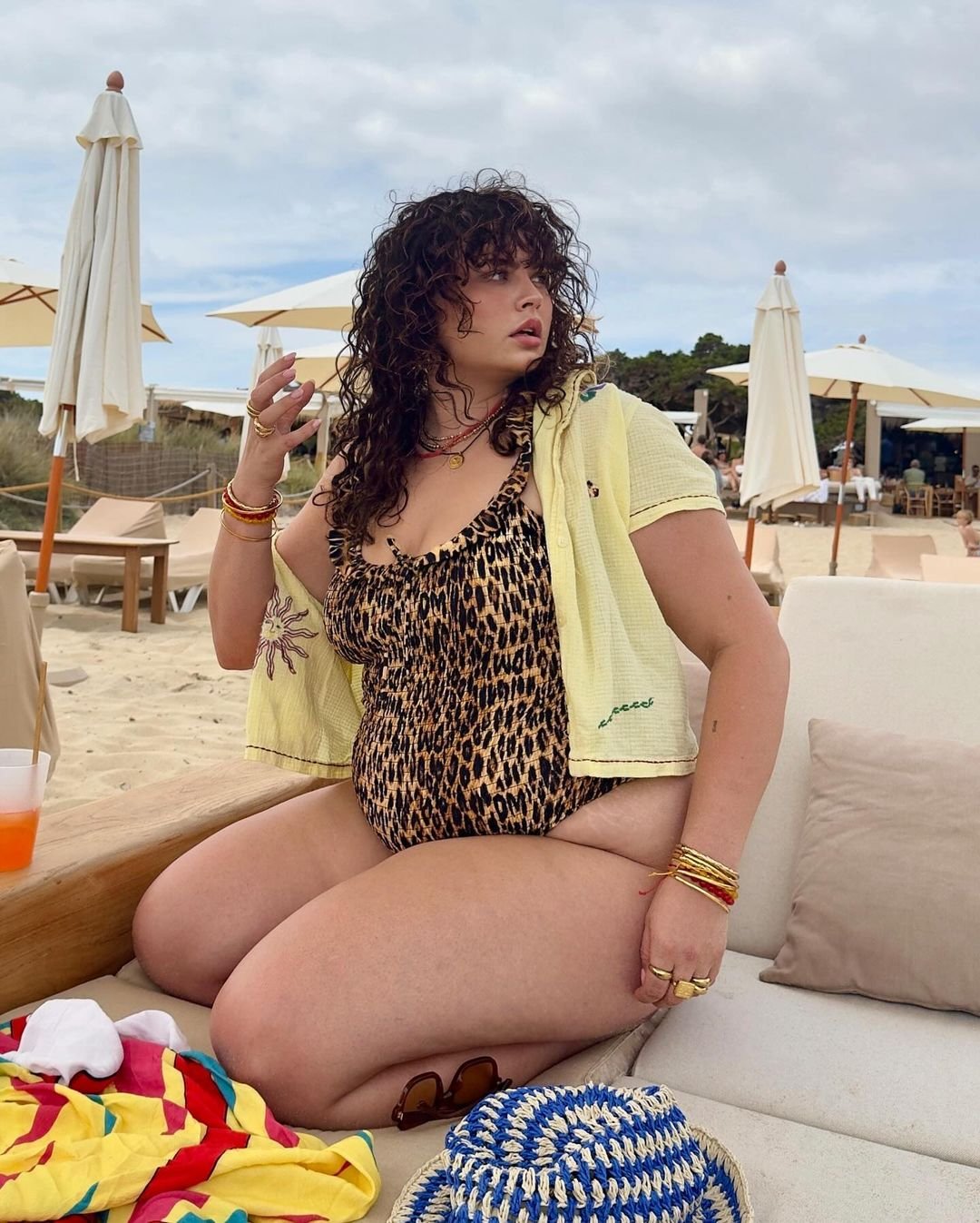 Mulher na praia usando maiô de estampa animal print, sobreposto por uma camisa leve amarela. Acompanha acessórios modernos, como pulseiras coloridas e colares. Tendência focalizada em conforto com estilo descontraído e vibrante para climas quentes.