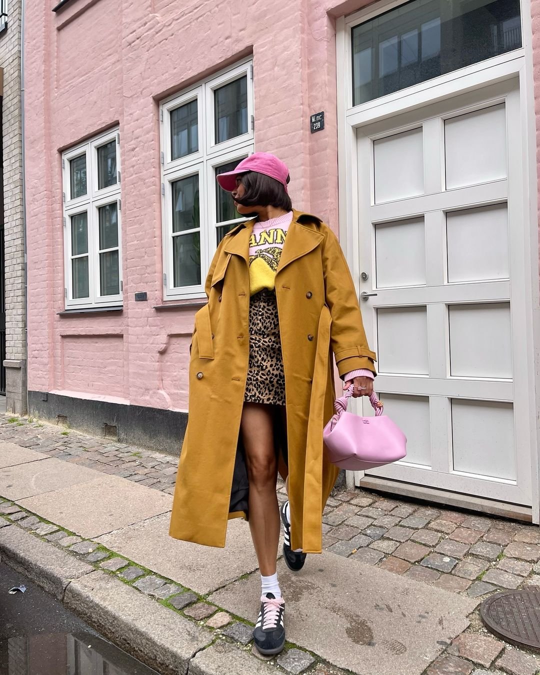 Pessoa vestindo um trench coat amarelo, camiseta com estampa de leopardo, tênis preto com meias brancas, boné rosa e bolsa rosa. Tendência de moda urbana e color block, misturando estampas animais com cores vibrantes. Cabelo ondulado e solto.