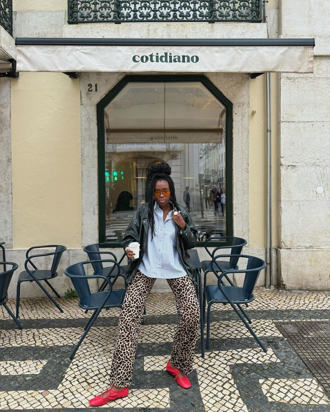 Pessoa vestindo roupa de oncinha, blusa branca, jaqueta de couro preta e sapatos vermelhos. Óculos de sol laranjas como acessório. O visual mescla ousadia e estilo casual, destacando tendências modernas e urbanas.