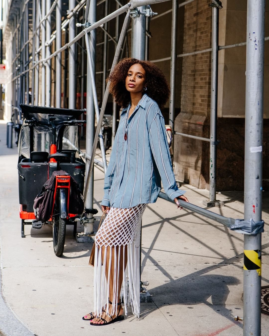 Pessoa vestida com camisa listrada azul oversized e saia de crochê branca com franjas longas. Completa o look com sandálias de tiras e acessório no pescoço. Estilo urbano e boho, misturando peças grandes com texturas artesanais.