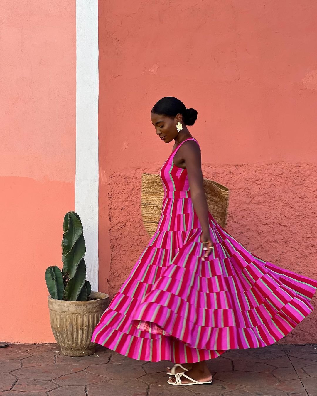 Imagem de mulher vestindo um vestido longo, listrado em tons de rosa e vermelho. A peça possui um design rodado e volumoso. Ela complementa o look com sandálias brancas e brincos grandes, destacando-se a tendência de acessórios chamativos.