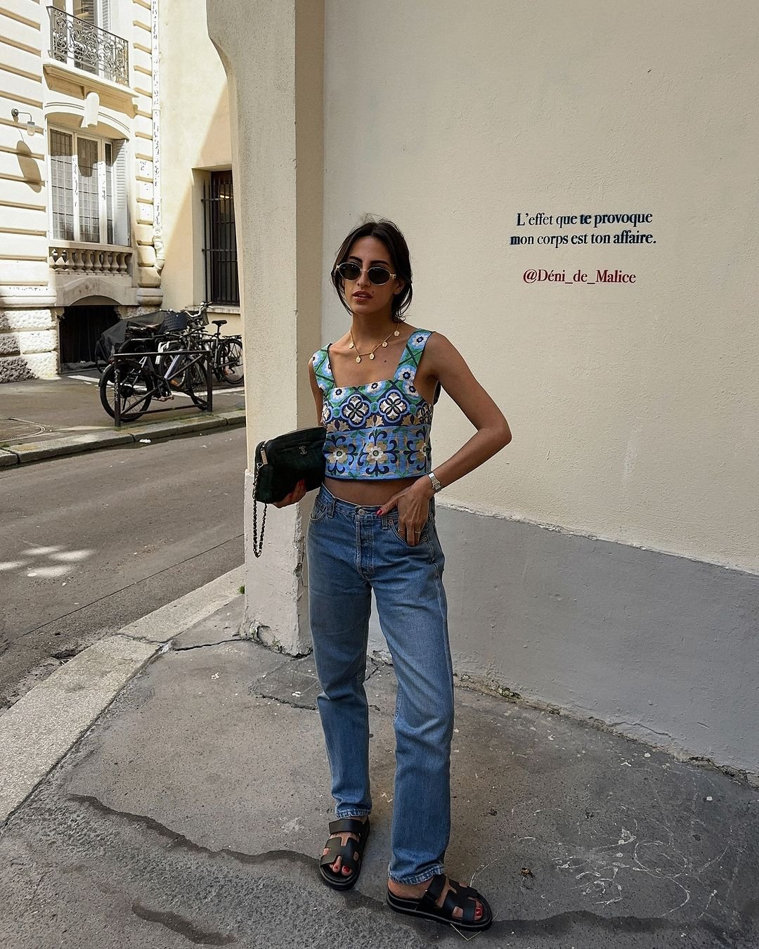 Mulher vestindo top cropped estampado colorido, jeans de cintura alta, sandálias pretas e óculos escuros, segurando uma bolsa preta pequena. O look é casual, moderno e adequado para o verão, refletindo tendências de moda urbana com um toque de descontração.