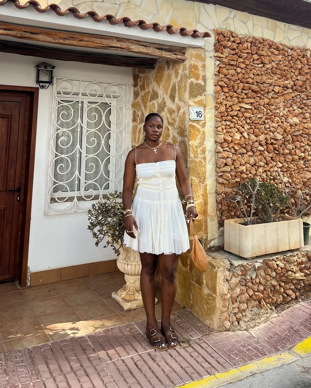 Mulher vestindo um vestido branco leve e esvoaçante com alças finas, sandálias de tiras e acessórios dourados. Bolsa marrom casual. Visual fresco e descontraído, ideal para dias quentes e eventos ao ar livre. Combinação de tendências boho e clássica.