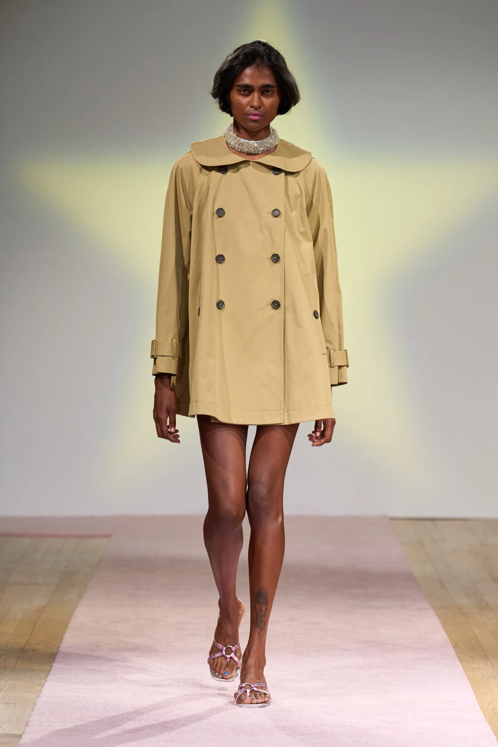 Modelo vestindo capa trench coat bege com abotoamento duplo, tendência primavera/verão. Look complementado por sandálias de salto baixo e colar volumoso. Estilo sofisticado e minimalista. Passarela e moda contemporânea destacam elegância casual.