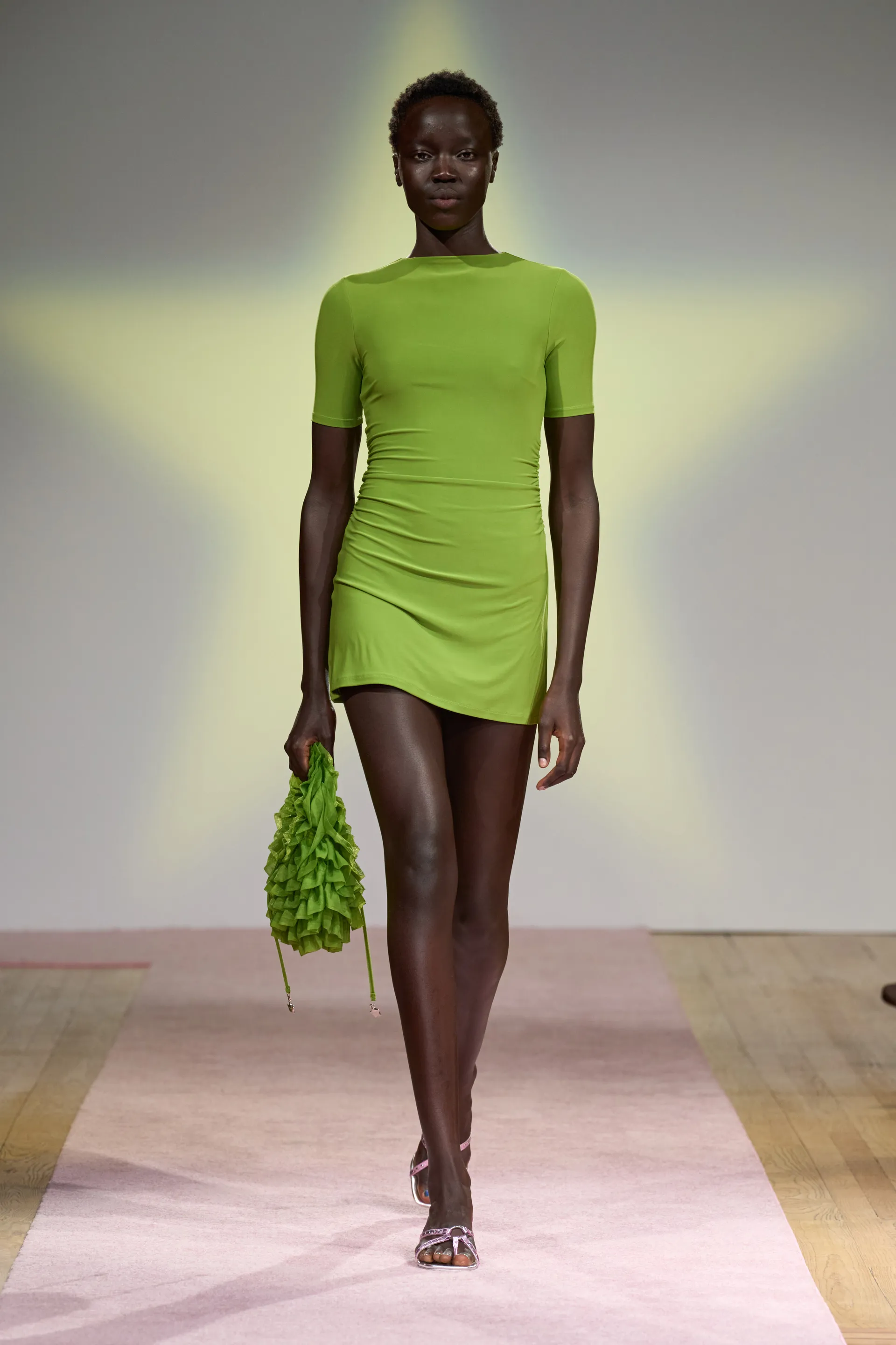 Modelo usa vestido curto verde com mangas curtas, bolsa com babados da mesma cor e sandálias de salto finas. Visual elegante e moderno reflete tendências de moda minimalista e monocromática para eventos e passarelas.