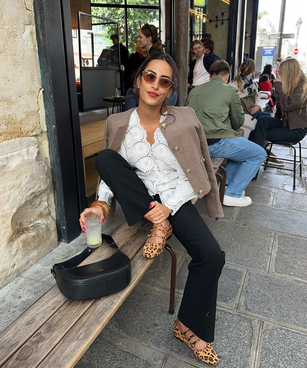 Mulher sentada em um café ao ar livre, vestindo uma camisa de renda branca, blazer marrom, calça preta e sapatilhas com estampa de leopardo. Acessórios incluem óculos de sol e bolsa preta. Look mistura elementos clássicos com trendy animal print.