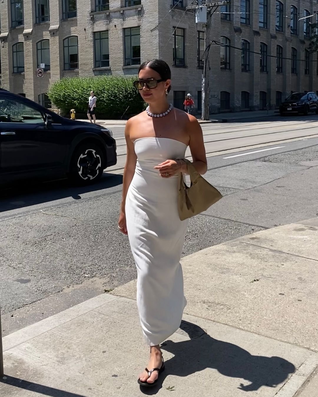 Mulher com vestido branco tomara-que-caia, sandálias pretas e bolsa bege. Usa óculos escuros grandes e colar de pérolas. Look minimalista e elegante, perfeito para dias quentes. Tendências de acessórios oversized e estilo clean.