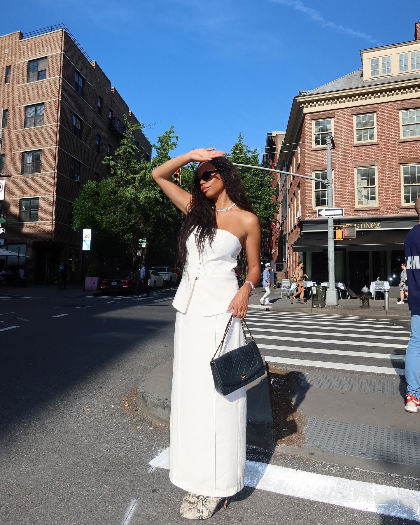 Rapariga com look elegante de verão: vestido longo branco strapless com corte moderno, bolsa preta de corrente dourada e óculos de sol grandes. Combinações minimalistas e sofisticadas influenciadas pelas últimas tendências de moda urbana.