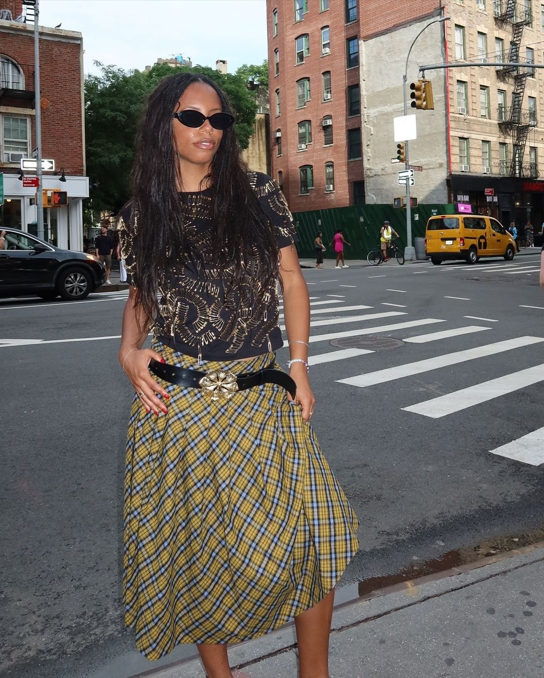 Mulher com estilo urbano, usando óculos de sol modernos, camiseta preta estampada com detalhes dourados, saia xadrez amarela e azul, e cinto preto com fivela decorativa. Look combina tendências contemporâneas e peças clássicas. Rua movimentada ao fundo.