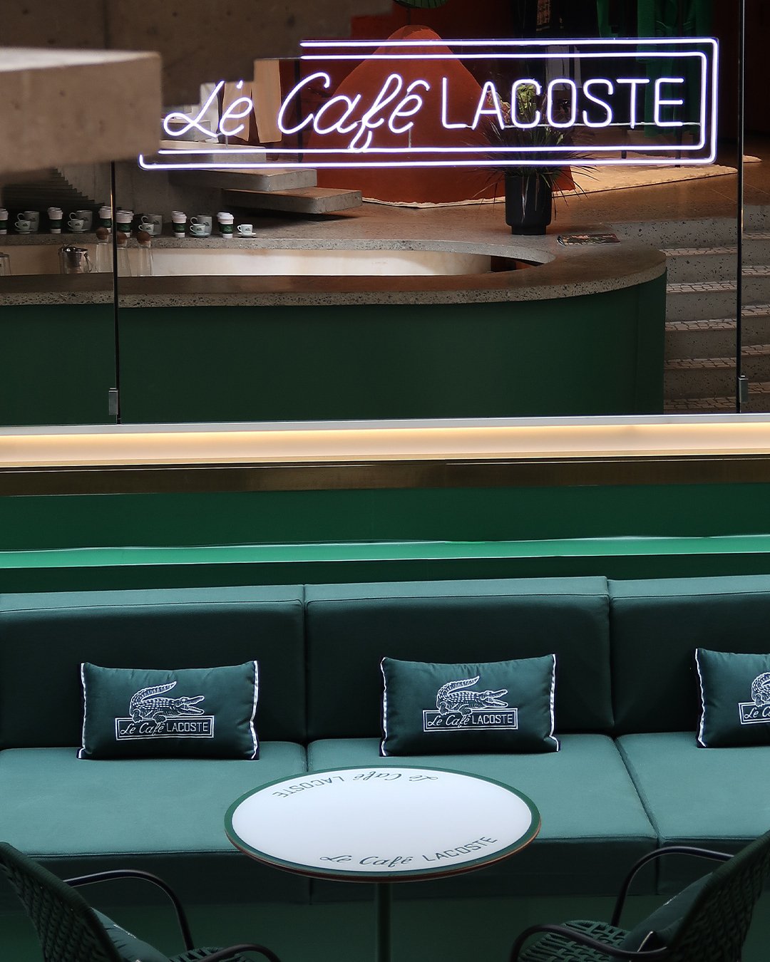 Imagem do Le Café Lacoste na NK Store, destacando um ambiente moderno e elegante com tons verdes, almofadas e decoração minimalista.