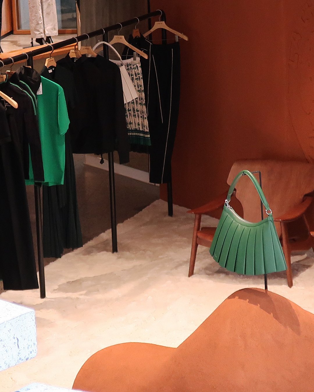 Loja de moda contemporânea com roupas sofisticadas em tons de preto, verde e estampas elegantes. Destaque para combinações de blusas, saias e calças penduradas em cabides. NK Store.