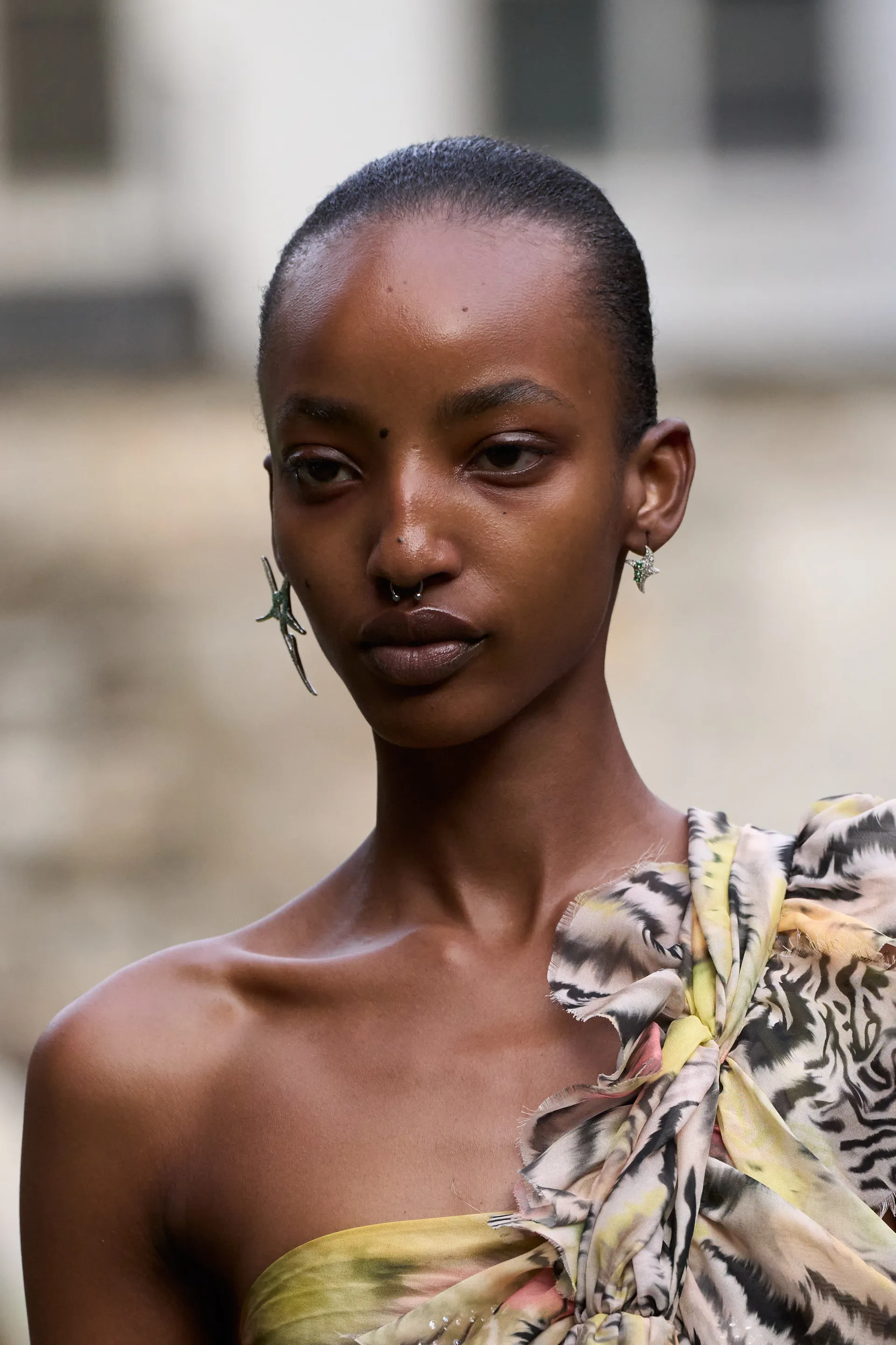 Modelo com penteado minimalista, vestindo um top de um ombro só com estampa animal print em tons de amarelo e cinza. Acessórios incluem piercing no septo e brincos pequenos e detalhados. NYFW 2025.