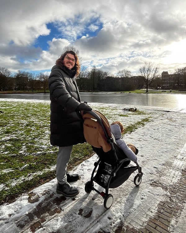 Homem usando um casaco longo e acolchoado preto, luvas pretas e calça clara empurrando um carrinho de bebê. Look de inverno, apropriado para clima frio com neve. Botas escuras completam o visual funcional e confortável.