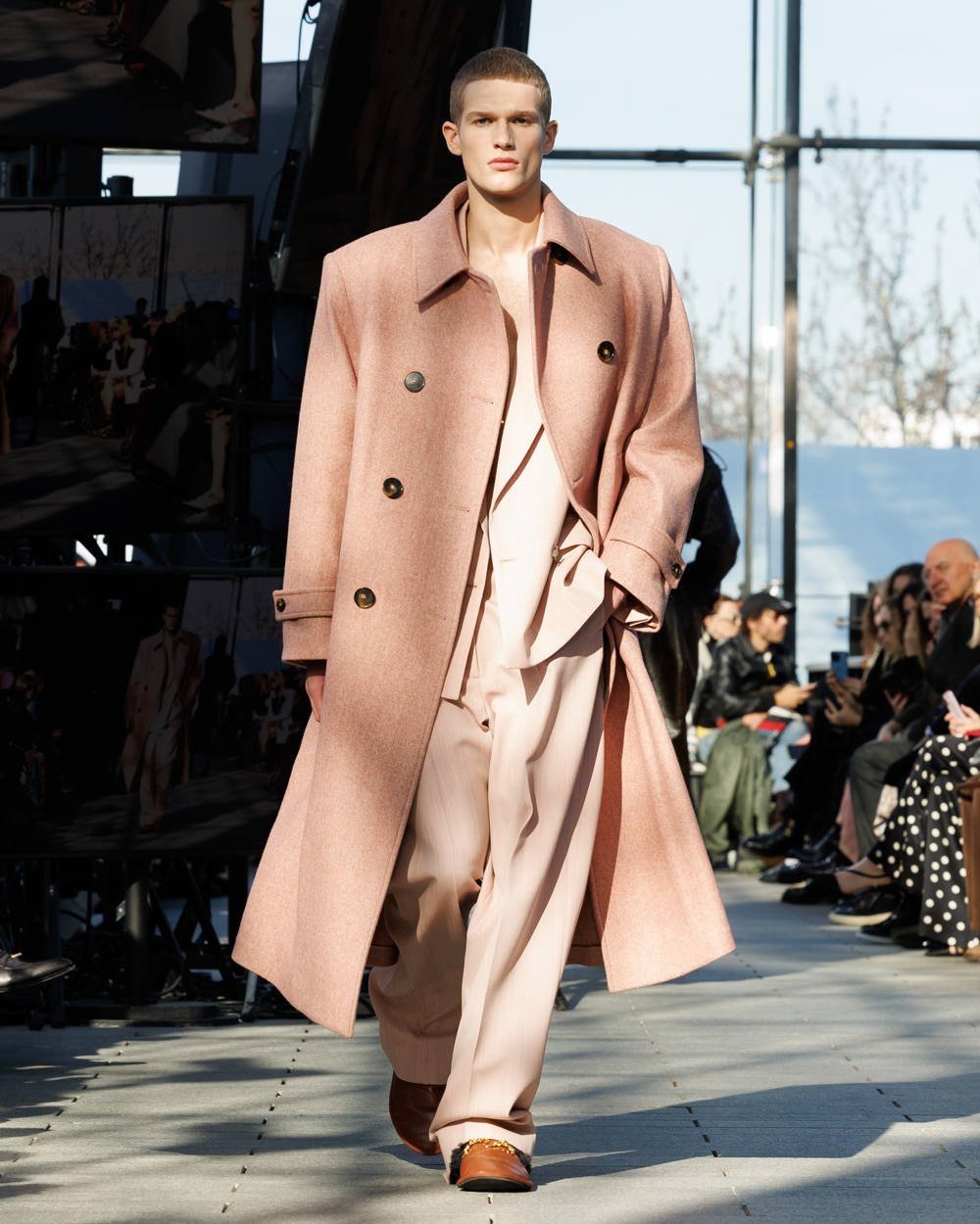 Desfile de moda masculina. Modelo veste sobretudo rosa em alfaiataria com abotoamento duplo, sobre conjunto de calça e blazer em tom pastel. Look combina elegância vintage e tendências de moda contemporânea, destacando tons suaves e corte oversized.