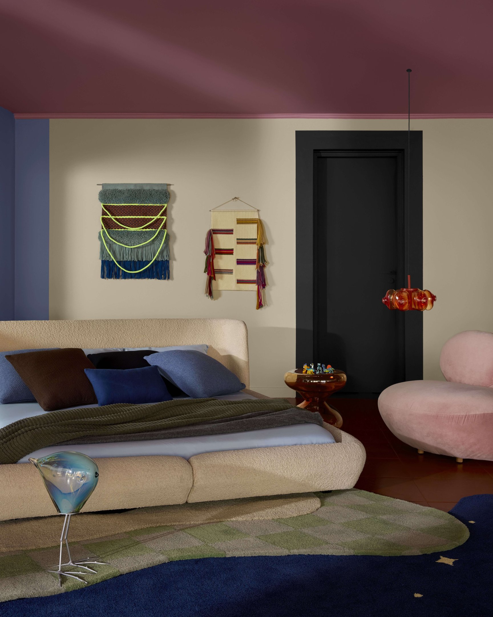 Quarto moderno e aconchegante com cama baixa bege, almofadas em tons de azul e marrom, tapeçaria vibrante na parede, poltrona rosa, e decoração com pendente laranja. Combinação de cores quentes e frias, unindo estilos contemporâneos e aconchegantes.