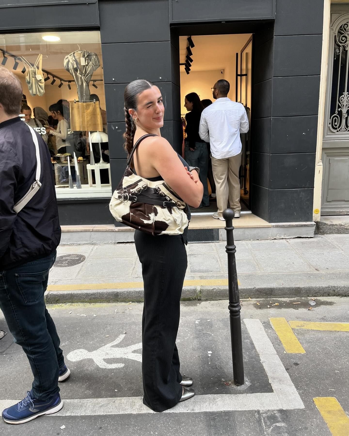 Mulher com estilo moderno veste calça preta de alfaiataria, top preto e blusa amarrada na cintura. Acessório: bolsa com estampa de vaca. Sapatos metálicos e cabelo em trança completam o look, que mistura tendências urbanas e sofisticadas.