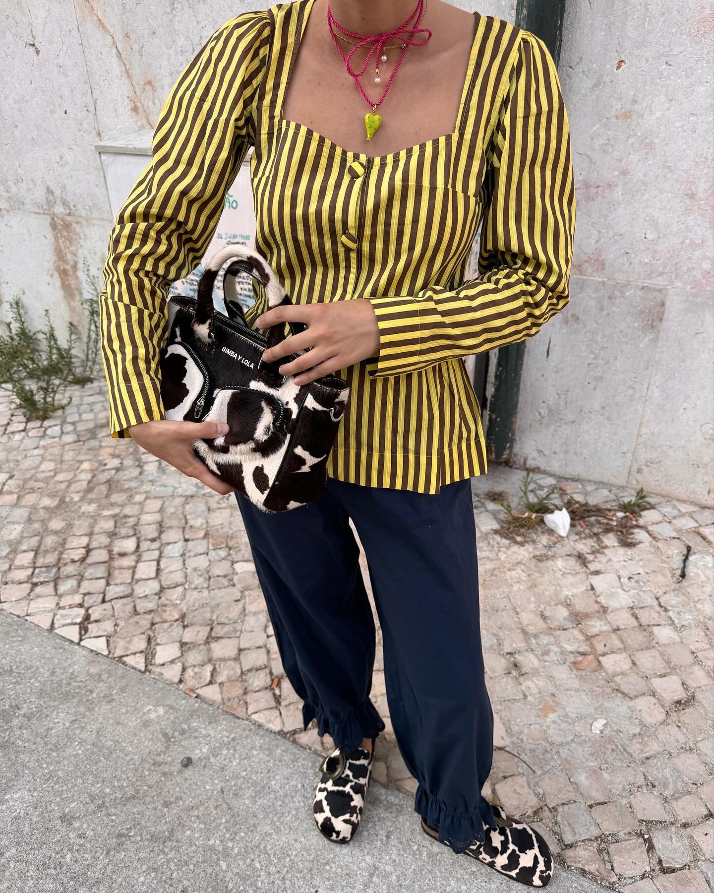 Pessoa vestindo uma blusa listrada em amarelo e preto, calça azul marinho com detalhes nas barras e acessórios de moda estampados em animal print. Carrega uma bolsa com estampa de vaca e usa sapatos combinando, completando o look com colares modernos e vibrantes.