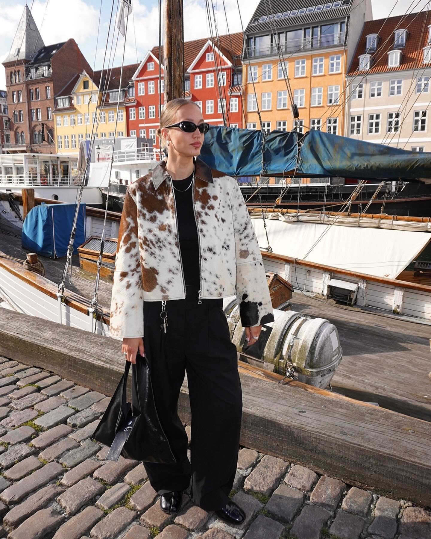 Pessoa usando jaqueta estampada em padrão animal, óculos escuros estilizados, calça preta solta e sapato preto clássico. Carrega uma bolsa preta triangular. O look é urbano e moderno, combinando tendências de moda contemporâneas.