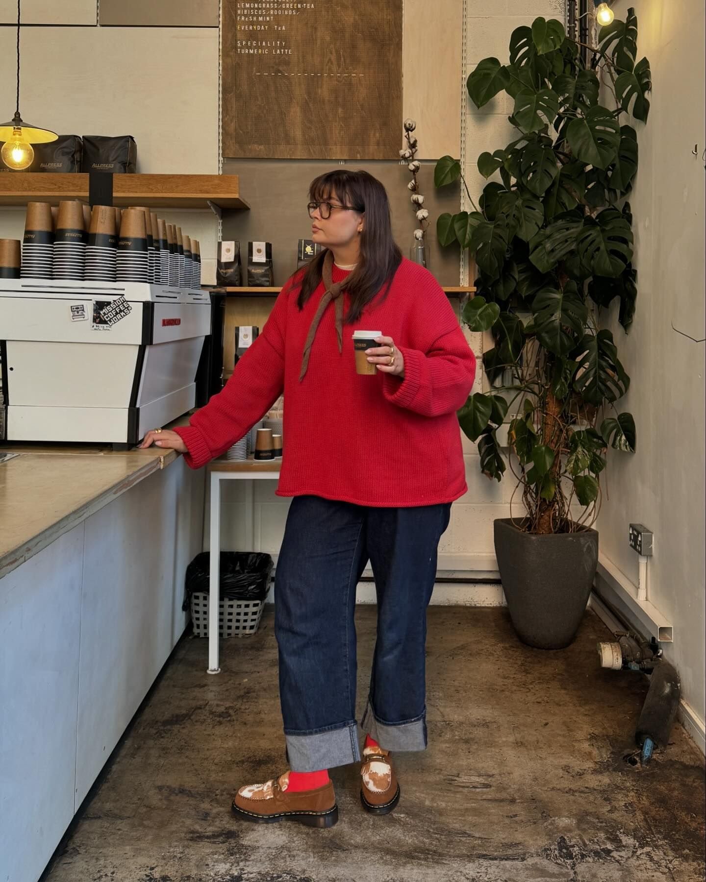 Pessoa em uma cafeteria, usando suéter vermelho, jeans e sapatos estampados. Ela segura um copo de café.