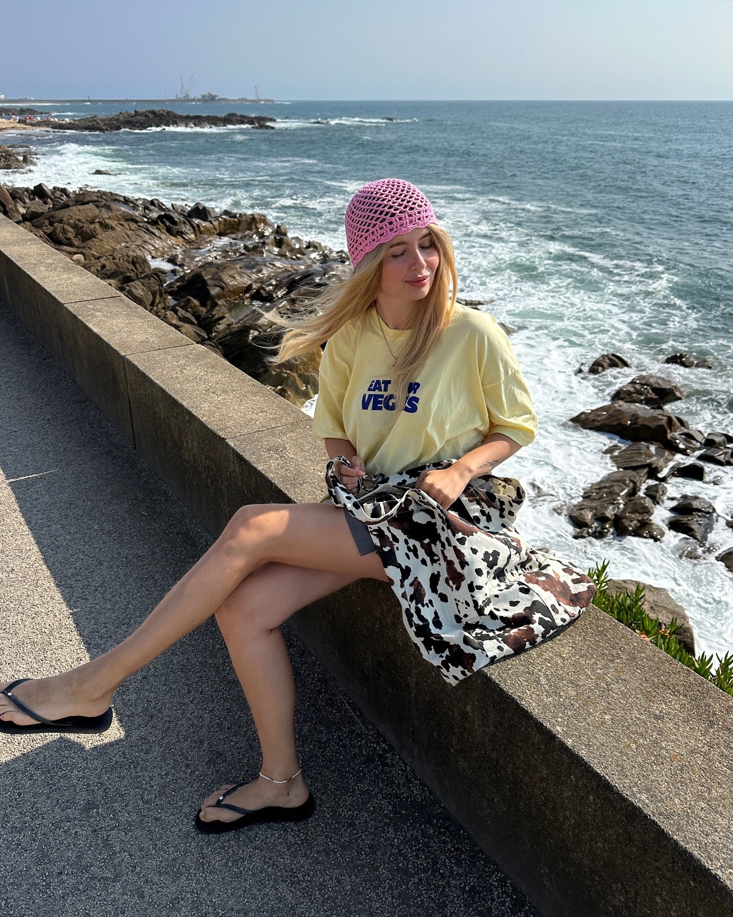 Imagem mostrando uma jovem sentada à beira-mar, vestindo camiseta amarela estampada, chapéu de crochê rosa e sandálias pretas. Bolsa com estampa de vaca complementa o visual descontraído, refletindo tendências de moda casual e confortável para o verão.