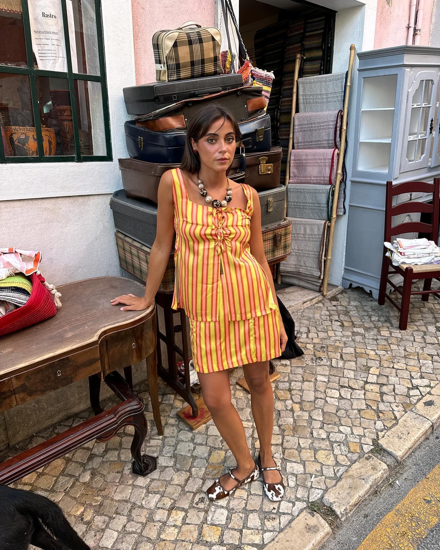 Mulher vestindo um vestido de verão listrado em amarelo e vermelho, com alças finas e franzido no busto. Ela usa uma sandália com estampa animal print e um colar grande. Cenário de rua com malas empilhadas, móveis antigos e objetos vintage ao fundo.