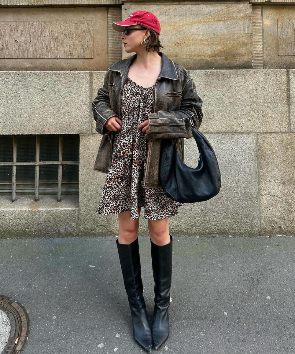 Pessoa usando vestido de estampa animal print, jaqueta oversized de couro, botas de cano alto e bico fino pretas. Complementa o look com boné vermelho, óculos de sol, brincos grandes dourados e bolsa preta. Estilo urbano e vintage.