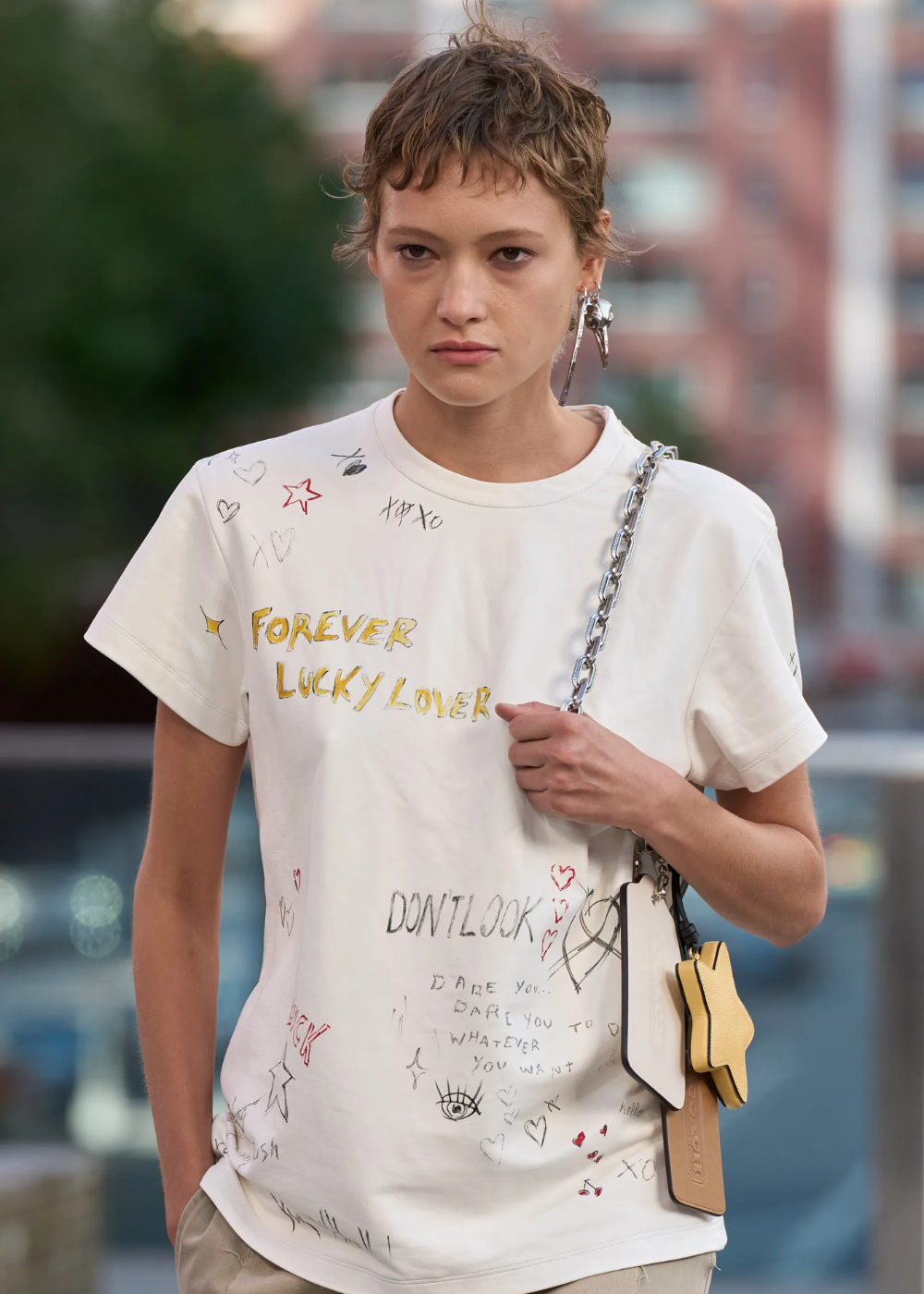 Pessoa vestida com camiseta branca estilo grafitada, com desenhos e frases, combinada com uma bolsa de alça de corrente prateada e acessórios modernos. O look casual destaca-se pelo visual jovem e despojado, ideal para quem busca estilo urbano e descontraído.