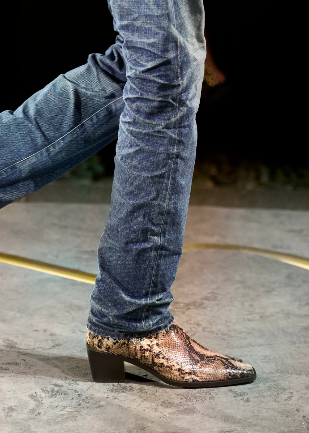 Pessoa usando jeans slim fit azul escuro combinado com botas de couro estampadas com padrão de cobra. As botas têm salto baixo, evidenciando um look moderno e ousado, misturando casual com elementos sofisticados. Moda urbana e contemporânea.