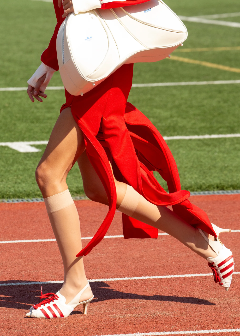 Pessoa vestindo um vestido vermelho vibrante com recortes, combinando com meias beges e sandálias de salto baixo em estilo esportivo com listras vermelhas no campo de uma pista de atletismo. Estilo moderno e ousado, misturando elementos esportivos e elegantes.