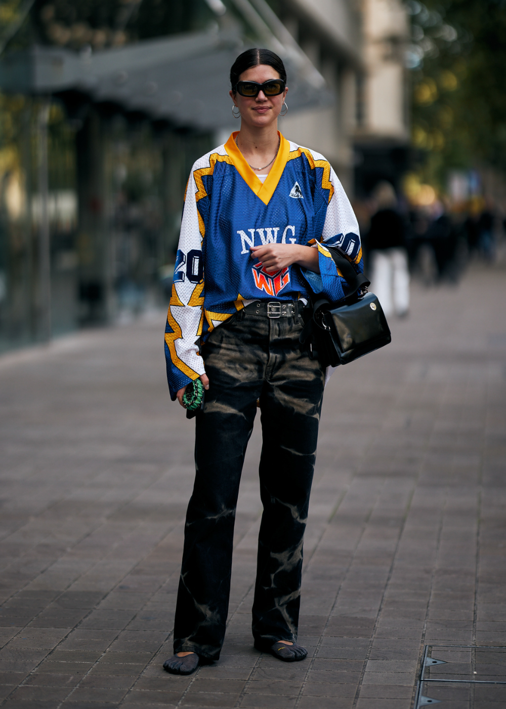 Pessoa vestindo uma camisa de hóquei azul e amarela com estampa, calças jeans largas e desbotadas, sapatos escuros e bolsa preta. Óculos de sol grandes e cabelo preso completam o visual urbano e moderno. Estilo casual e despojado com influência esportiva.