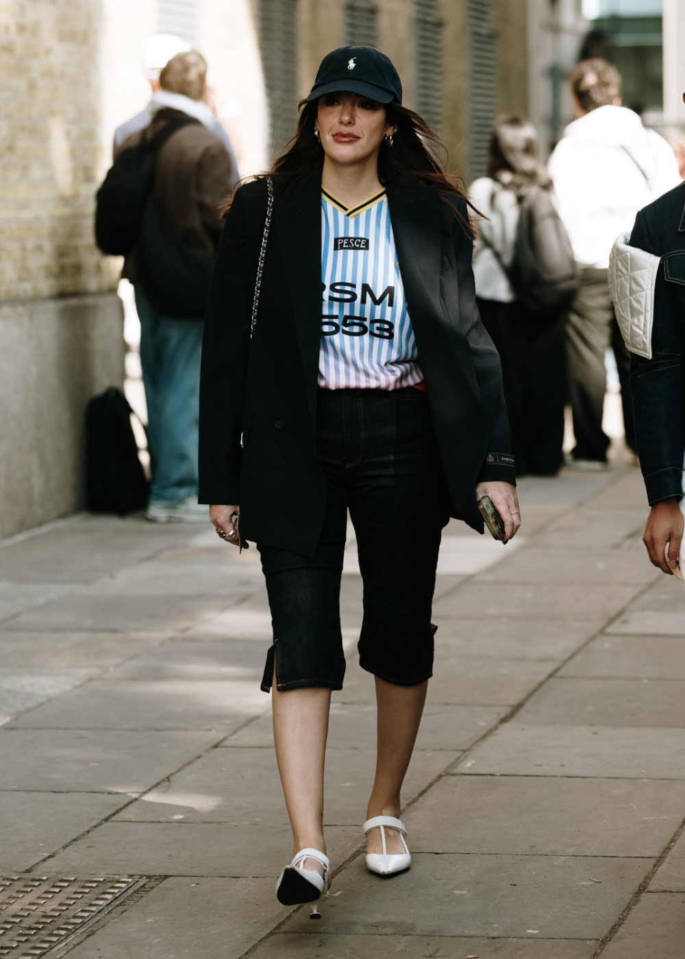 Mulher com look casual e estiloso usa t-shirt listrada em branco e azul com blazer preto, bermuda jeans escura e sapatilhas brancas. O toque esportivo é complementado por um boné preto. Tendências de moda urbana e casual chic.