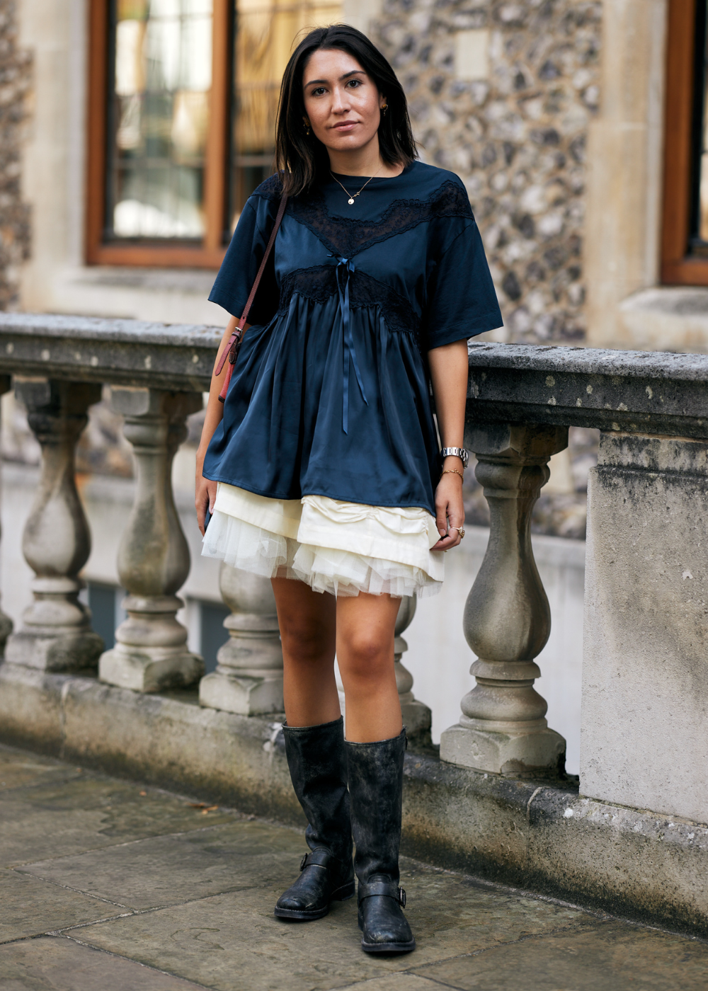 Mulher usando blusa de seda azul-marinho com renda e laço, saia com camadas de tule branco, e botas pretas de couro estilo motociclista. Look descontraído, misturando elegância e rebeldia, tendência boho chic e romantismo urbano. Ideal para destaque em moda de rua.