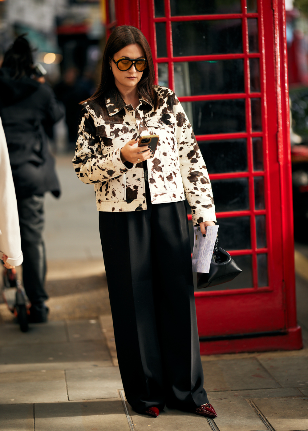 Mulher vestindo jaqueta com estampa de vaca, óculos de sol grandes e calça preta fluida. Estilo sofisticado com toque casual, combinando estampa animal com acessórios modernos, destacando-se em ambiente urbano. Tendência atual de misturar padrões e conforto.