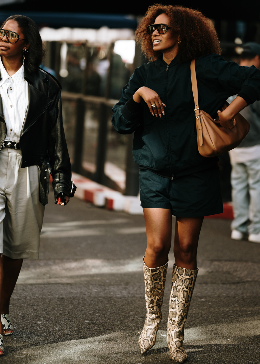 Duas mulheres na rua, uma com jaqueta de couro preta, camisa branca e shorts bege, a outra com jaqueta bomber preta, shorts esportivo preto e botas de cano alto com estampa de cobra. Ambas usam óculos de sol estilosos, refletindo tendências de moda urbana.