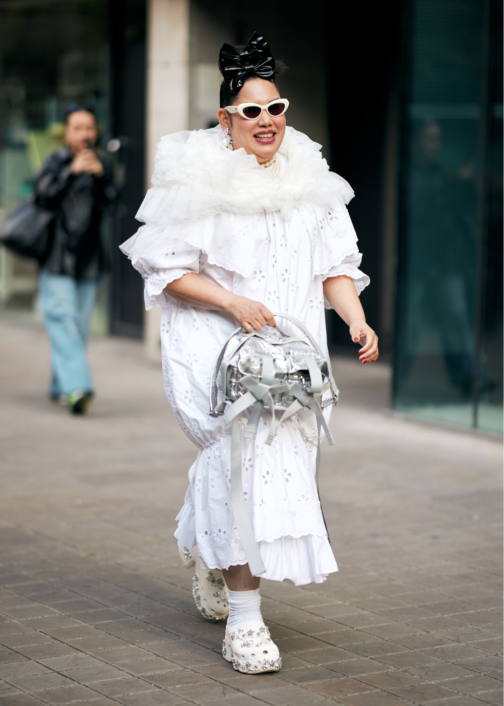 Pessoa com vestido branco rendado, babados volumosos e detalhes florais. Usa bolsa prateada futurista, óculos de sol brancos clássicos e sapatos plataforma brancos com pedrarias brilhantes. Laço grande preto na cabeça e meias brancas completam o look ousado.