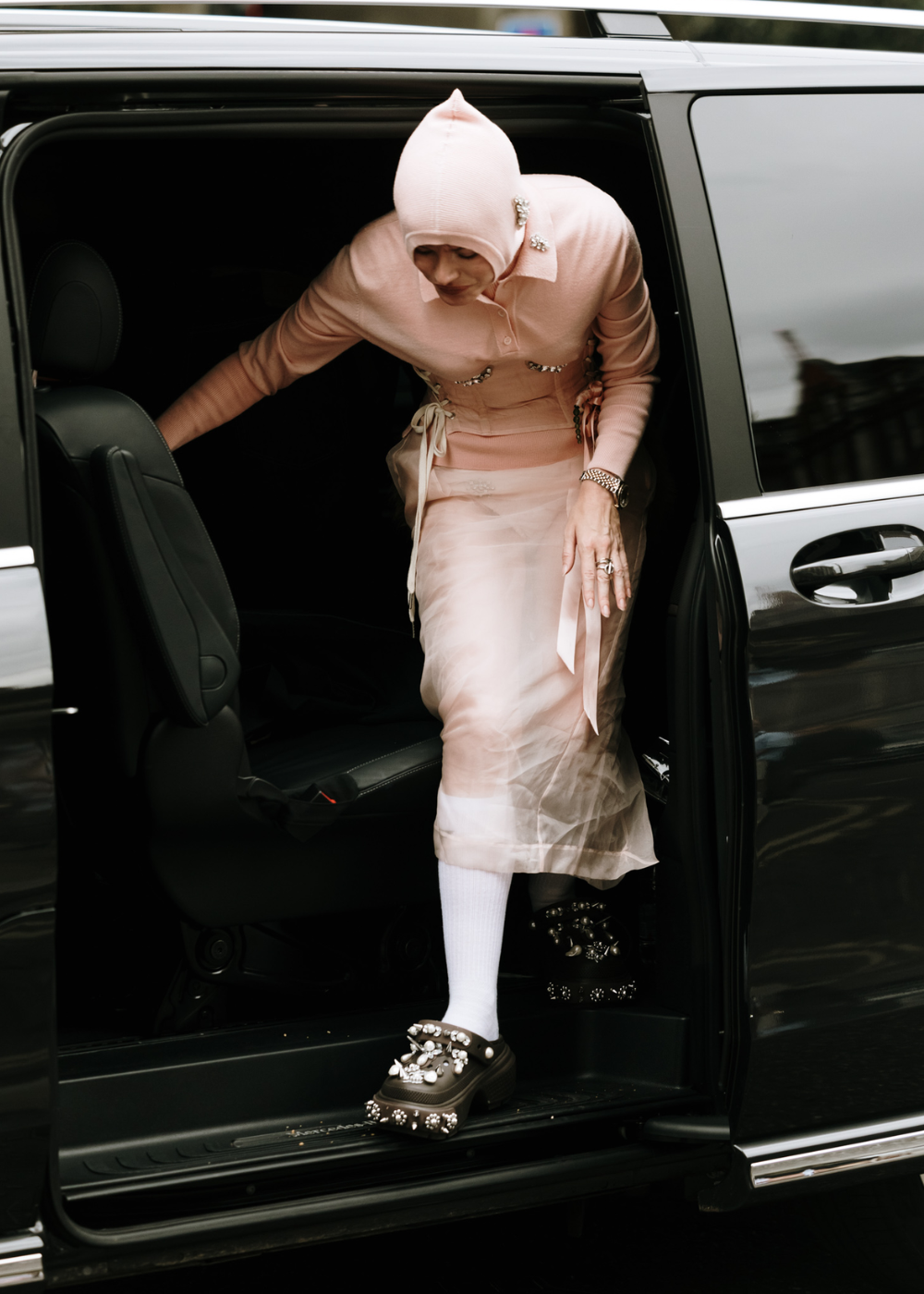 Pessoa saindo de um carro usando um conjunto rosa claro, com capuz e corset, saia transparente e meia branca. O look é complementado por sapatos plataforma decorados com pedrarias, refletindo uma tendência de estilo avant-garde e fashionista.