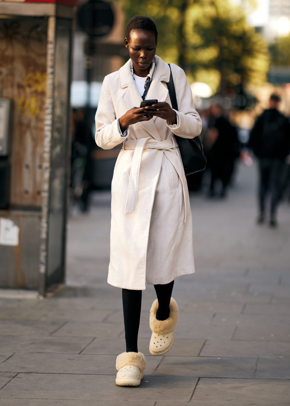 Pessoa caminhando na rua usando um sobretudo branco, calça preta e calçados tipo crocs com forro de pelúcia. Look é elegante e casual, combinando tendências modernas de conforto e estilo urbano. Acessório indispensável: bolsa preta discreta.