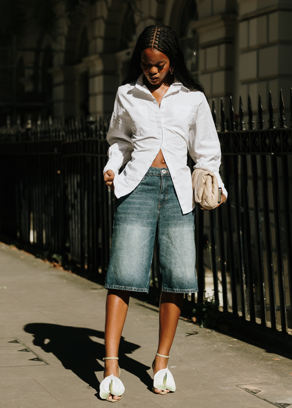 Mulher vestindo camisa branca e bermuda jeans no estilo casual chique. Ela complementa o look com uma bolsa clutch bege e sandálias de salto com detalhes florais. Tendência moderna e minimalista para um dia ensolarado na cidade.