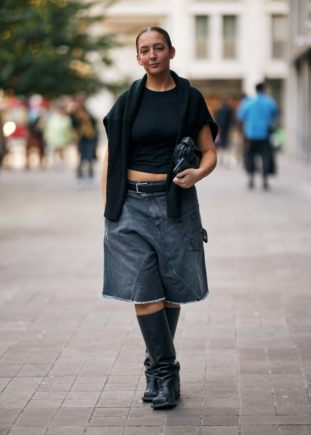 Mulher vestida com top preto cropped, colete de malha preto, bermuda jeans cinza e botas altas pretas. Segura uma clutch preta. Look urbano e casual com influência do estilo streetwear e tendência utilitária.