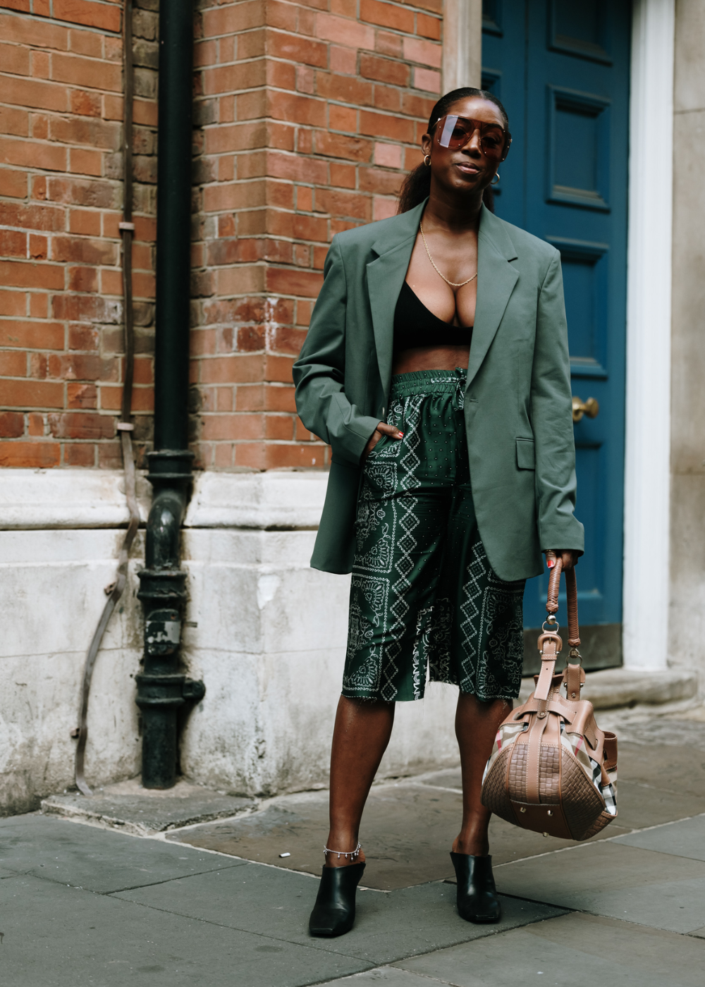 Mulher usando blazer oversized verde, top preto, bermuda estampada verde e sapatilhas pretas. Complementa o look com óculos de sol grandes e bolsa de couro marrom. Estilo moderno e casual com toques de elegância.