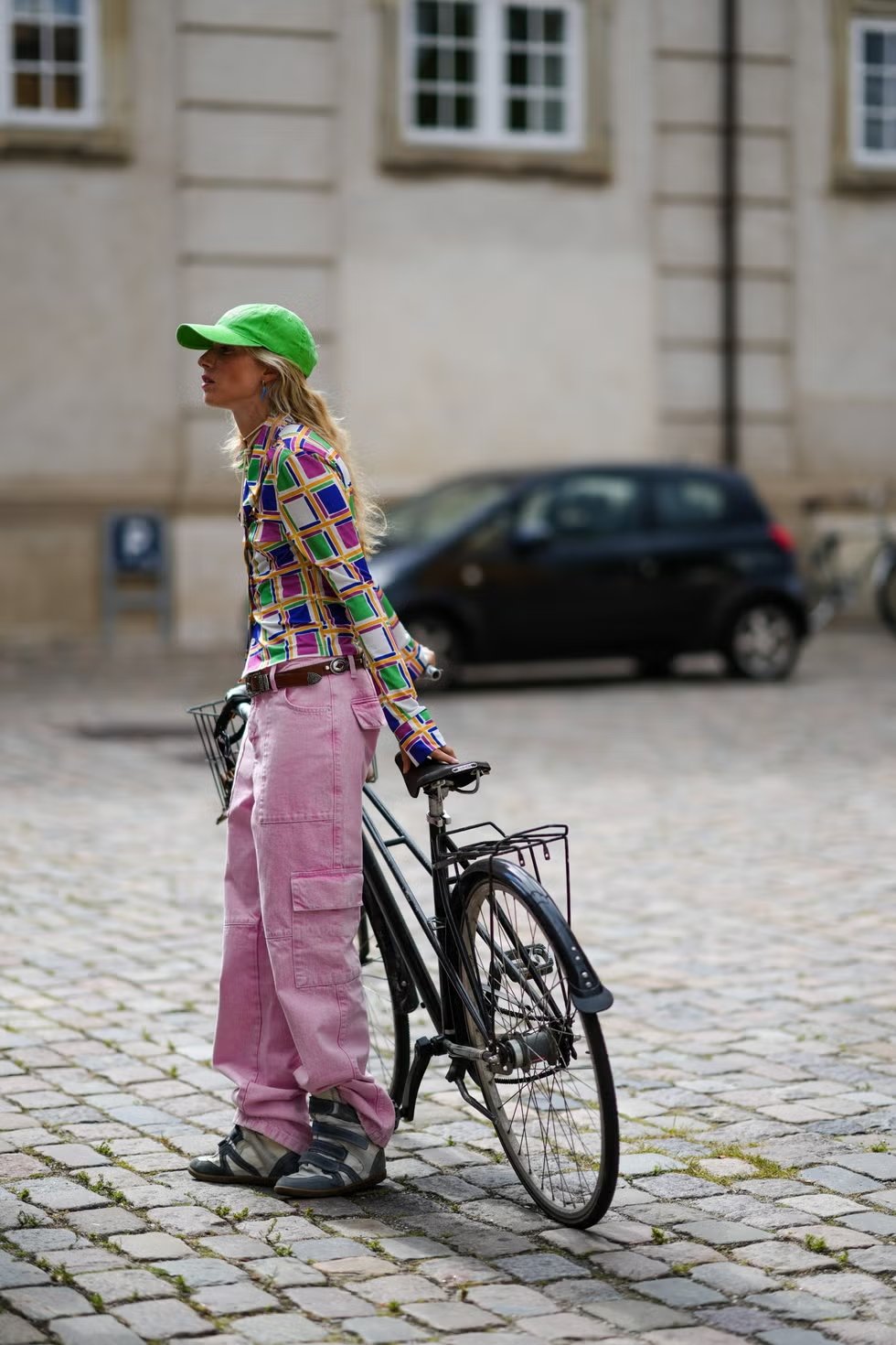 Pessoa vestindo uma camisa de manga longa com estampa colorida e geométrica, calça cargo rosa e boné verde. O visual é casual e moderno, destacando tendências vibrantes nos acessórios e nas peças com cores vivas e formas variadas. tênis de salto